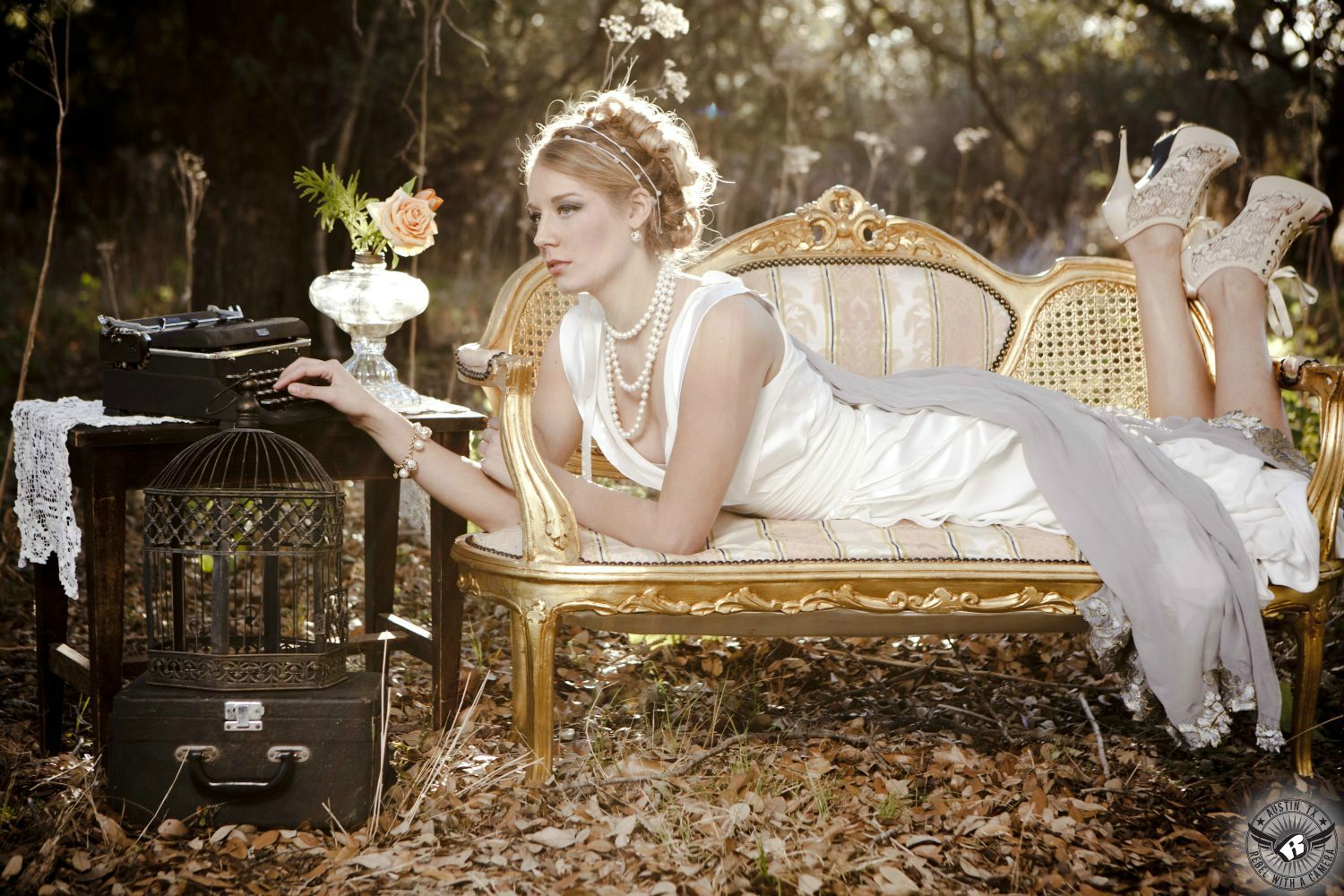 romantic blond wearing a long flowing vintage white dress and pearls and lacy heals laying on a vintage couch typing on an antique typewriter with an antique birdcage and suitcase in fall leaves